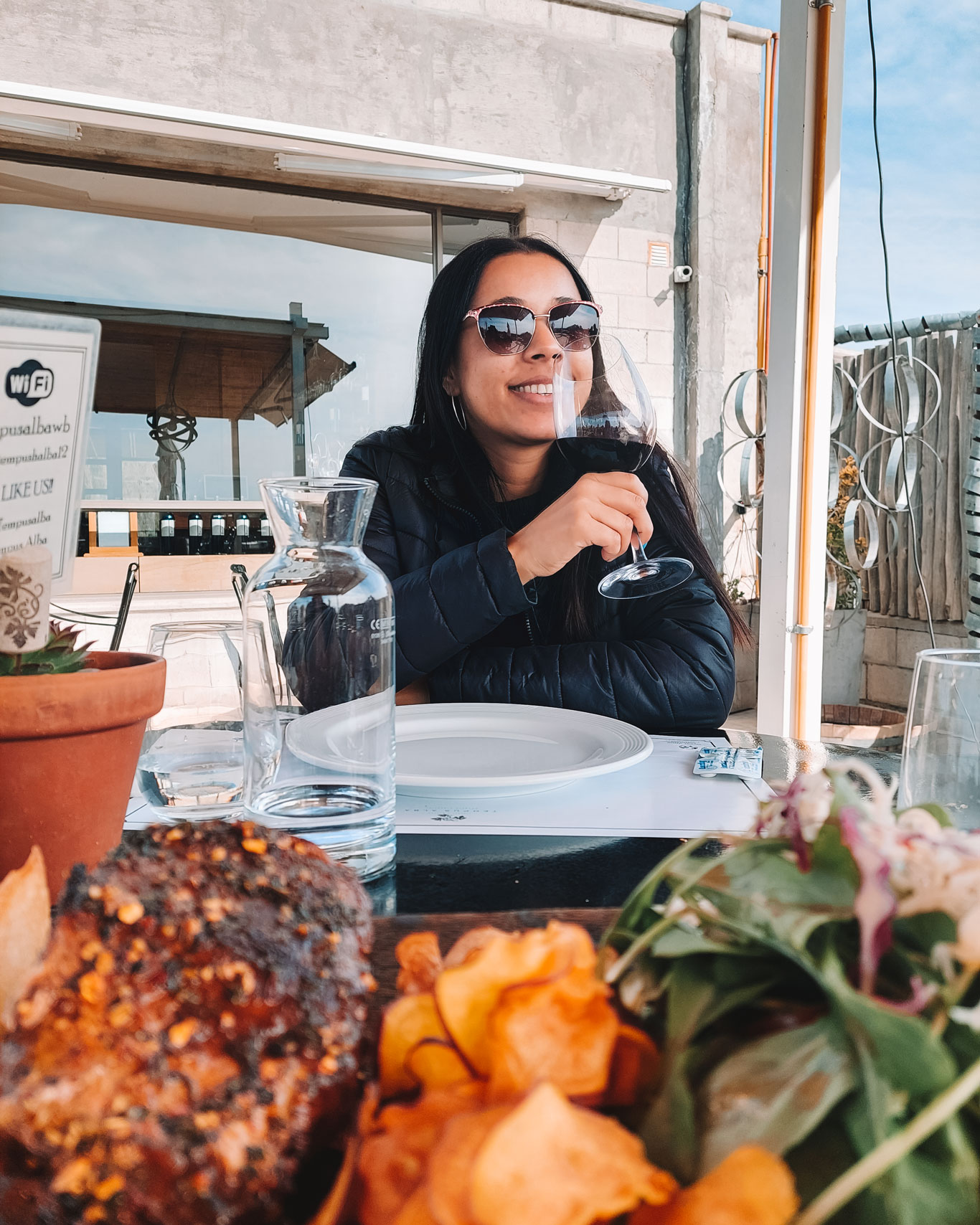 This image features a woman enjoying a glass of wine at an outdoor dining area of a vineyard. She is smiling, wearing sunglasses and a black puffer jacket, with a clear water jug and empty plates on the table. In the foreground, a platter with a grilled steak and crispy chips adds to the laid-back dining experience, underlined by a serene backdrop of vineyard scenery.