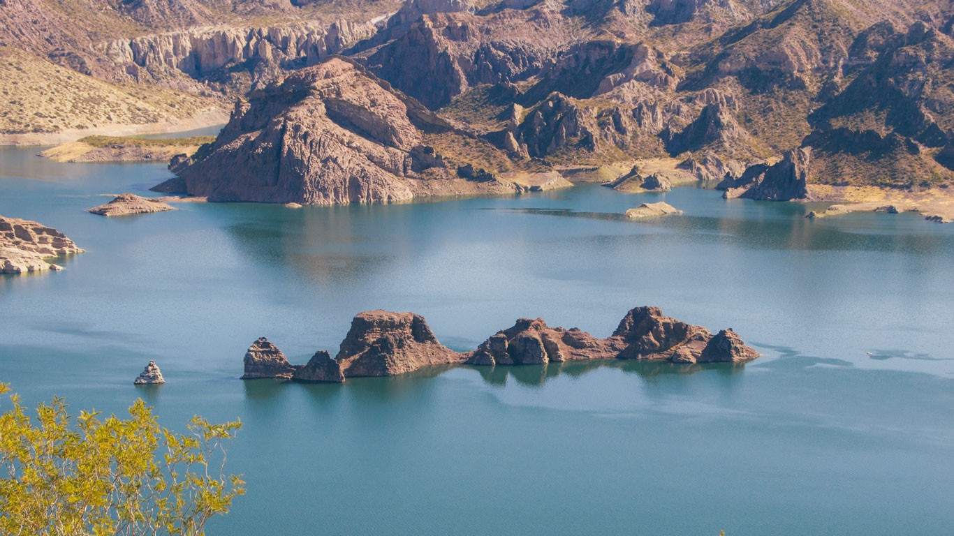 This image shows a serene view of a reservoir nestled among rugged mountains. The turquoise waters are calm, dotted with several rocky islands and outlined by craggy, brown mountain slopes. The foreground includes a glimpse of bright green foliage, enhancing the vibrant natural palette of the scene.