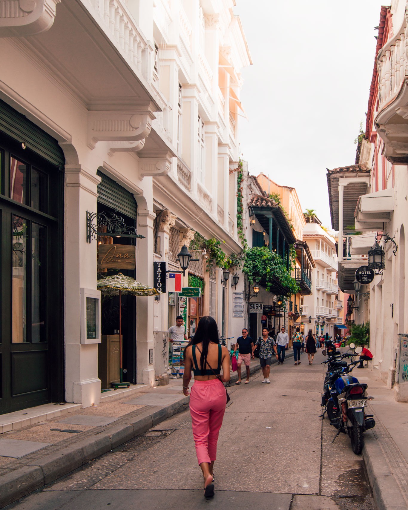 A woman walking through the narrow streets of downtown Cartagena, Colombia. She is seen from behind, wearing pink pants and a black blouse, with long black hair flowing casually as she walks. The streets are lined with white houses, featuring colorful doors and lush green plants adorning the balconies.