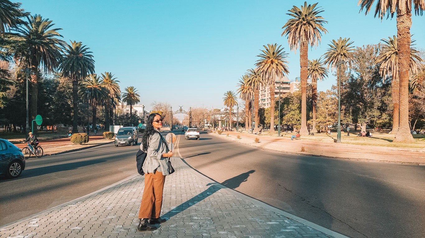 This image shows a person standing on a sunny street lined with tall palm trees, looking at the camera. They appear to be wearing a backpack and light, casual outdoor clothing. The street in Mendoza in September is relatively calm, with a few cars, cyclists and pedestrians visible.