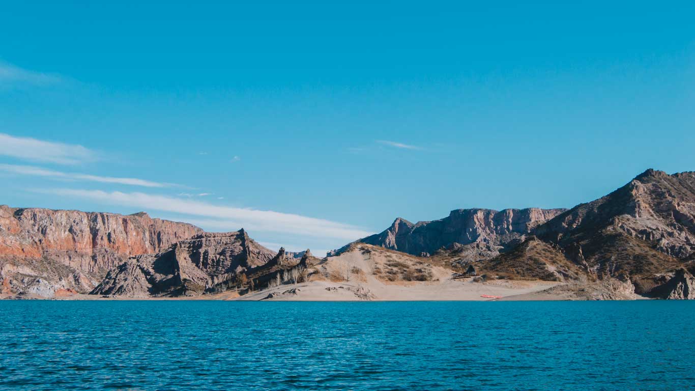 A panoramic view of a serene lake with azure waters in the foreground and rugged, rocky mountains in the background under a clear blue sky. The landscape features a mixture of barren rocky outcrops and small sandy beach areas, highlighting the natural beauty of the remote area.