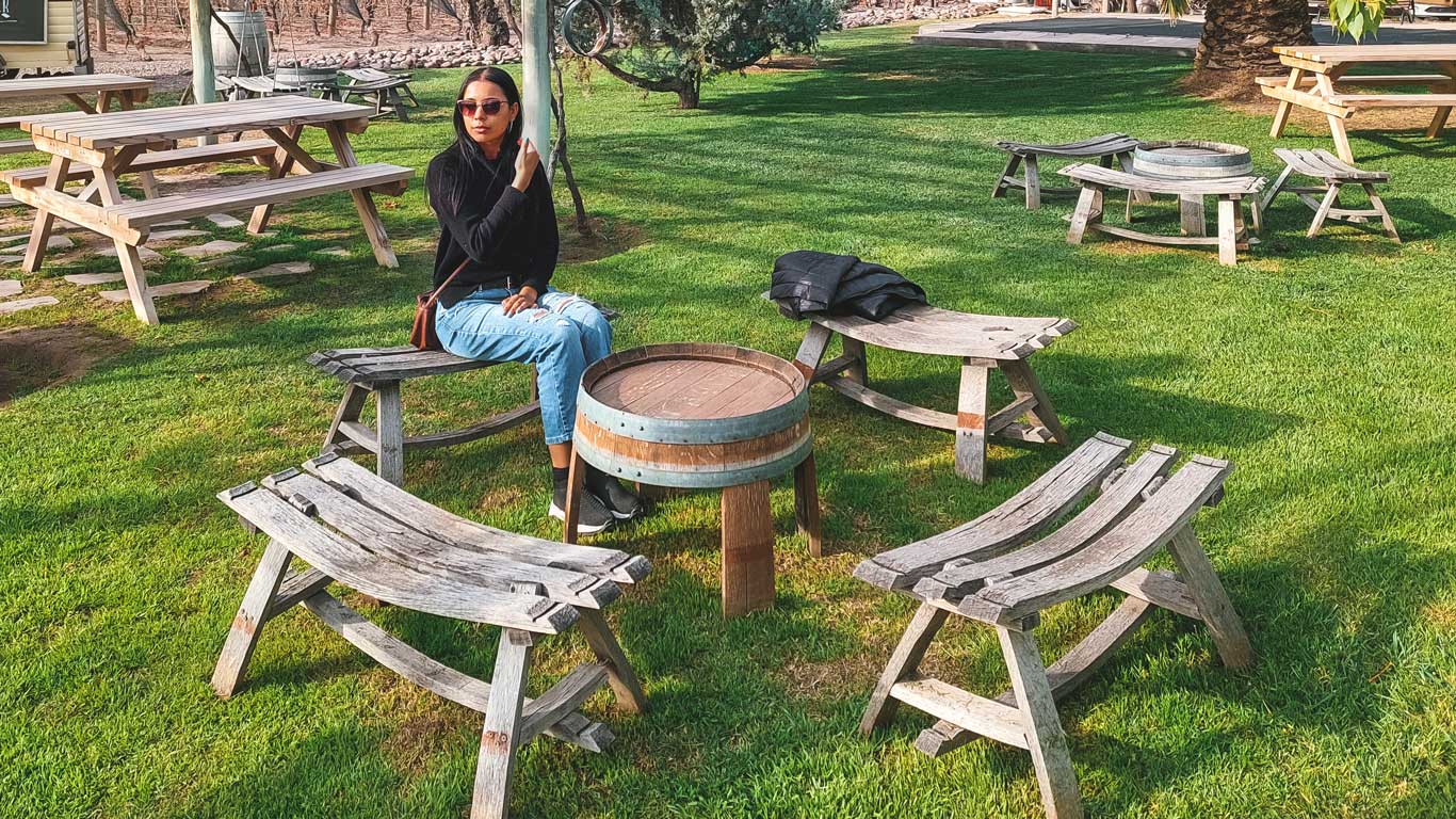 A woman wearing sunglasses and casual attire sits on a rustic wooden bench in a sunny vineyard garden. Surrounding her are circular tables crafted from wine barrels and additional wooden benches, set on a lush green lawn with trees softly swaying in the background.