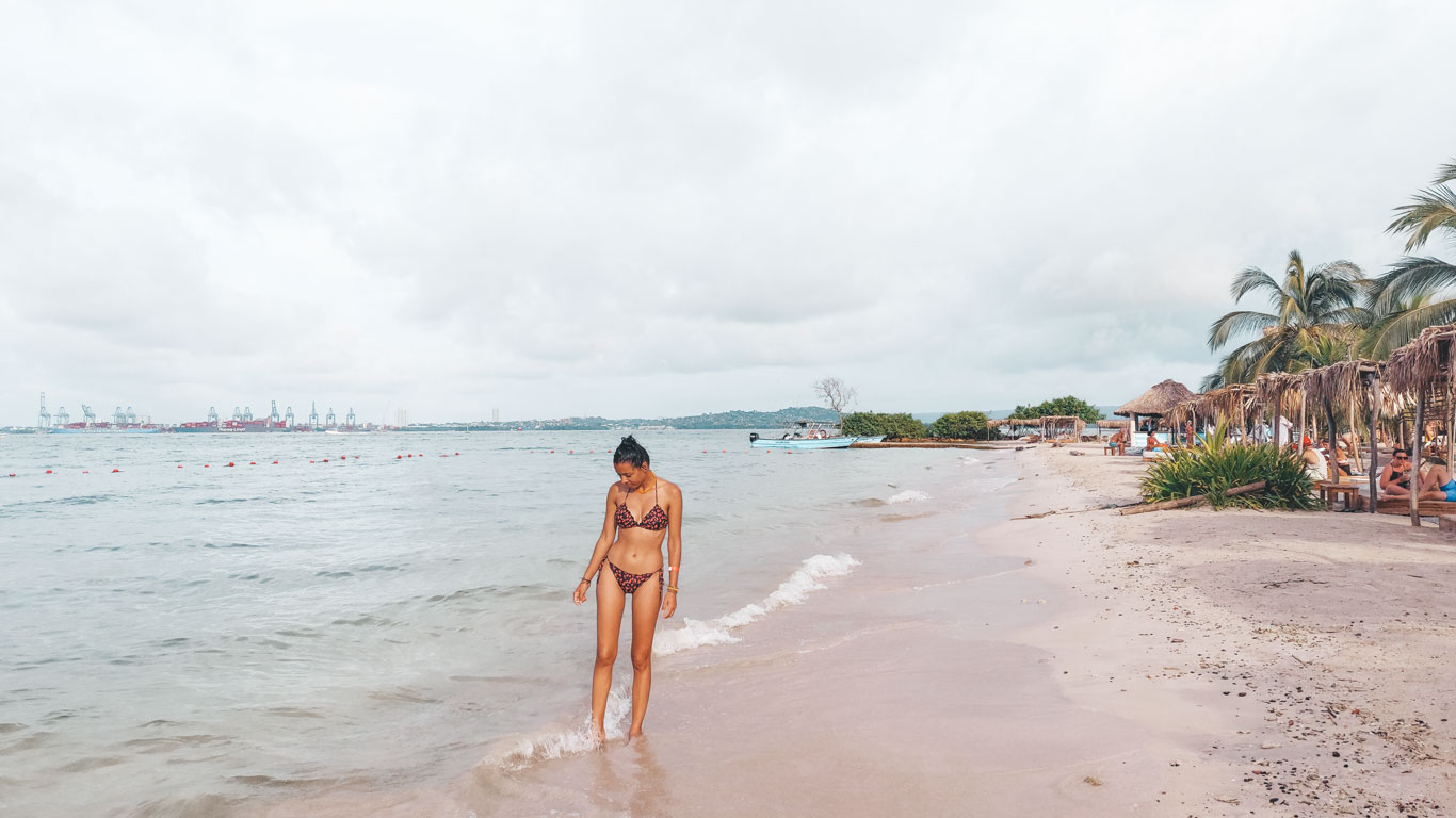 A woman wearing a purple bikini walking on the beach of Hotel Fenix Beach on Tierra Bomba Island. In the background, small tents with beach beds line the shore.