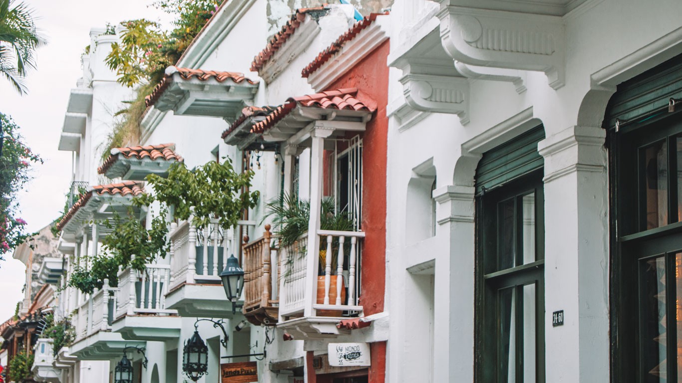 This image features a charming street view in Cartagena de Indias, showcasing a series of traditional colonial houses. The architecture is distinct with vibrant colors, ornate balconies, and classic tiled roofs. Lush greenery adds a touch of nature to the urban environment, creating a picturesque scene typical of this historic city.