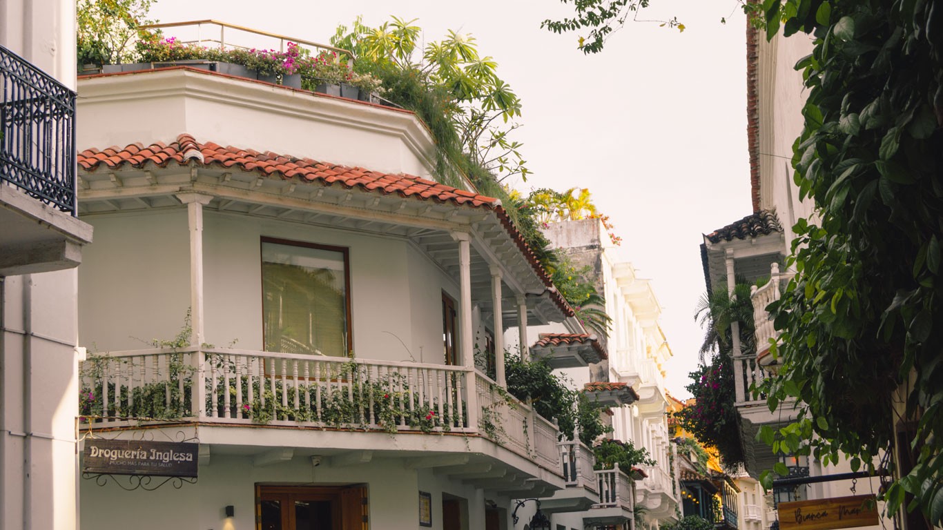 This image captures a serene street in Cartagena, lined with elegant colonial buildings adorned with lush rooftop gardens and ornate balconies. The architecture features white facades contrasted by dark blue details and wrought iron.