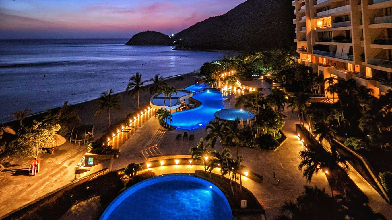 Night view of a beachfront resort in Bello Horizonte, Santa Marta, featuring several illuminated pools and palm trees around. In the background, you can see the beach and mountains under a sky painted by the sunset.