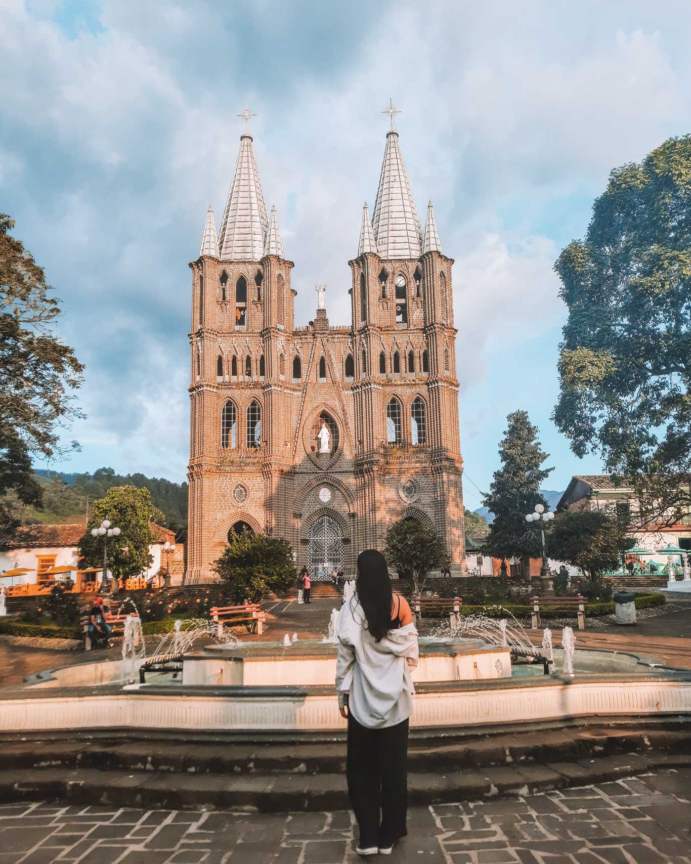 This image shows a woman standing in front of a beautiful brick cathedral with two tall spires, set against a backdrop of trees and mountains. A fountain is in the foreground, with water jets adding to the peaceful ambiance. The setting sun casts a warm light on the architecture, creating a serene atmosphere in what appears to be a central plaza in Jardín, Colombia.