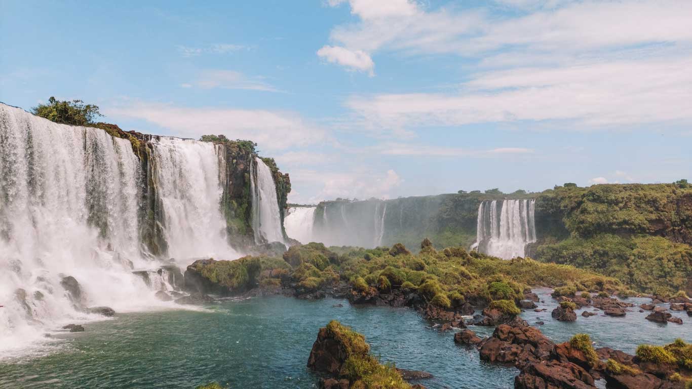 This image depicts the Iguazu Falls, a stunning series of waterfalls surrounded by lush greenery. The powerful cascades plunge into the Iguazu River, creating mist and a dramatic display of nature under a blue sky.