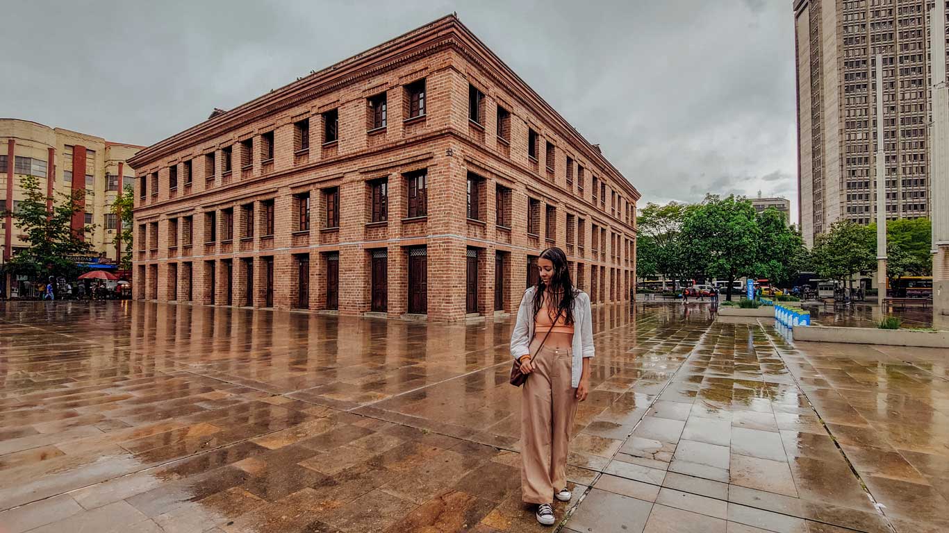 The image shows a rainy day scene in front of a historic brick building, with a woman standing in the foreground. The ground is wet and reflective, enhancing the moody atmosphere under a cloudy sky. Surrounding the scene are additional buildings and trees, creating an urban yet somewhat tranquil environment.