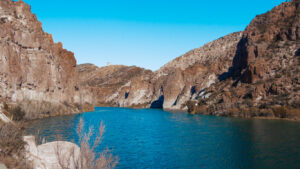 This image shows the Atuel Canyon in San Rafael, Mendoza. You can see towering rocky walls flanking a blue-water river, set in an arid, mountainous landscape under a clear sky. The canyon is a popular tourist destination due to its natural beauty and geological formations.