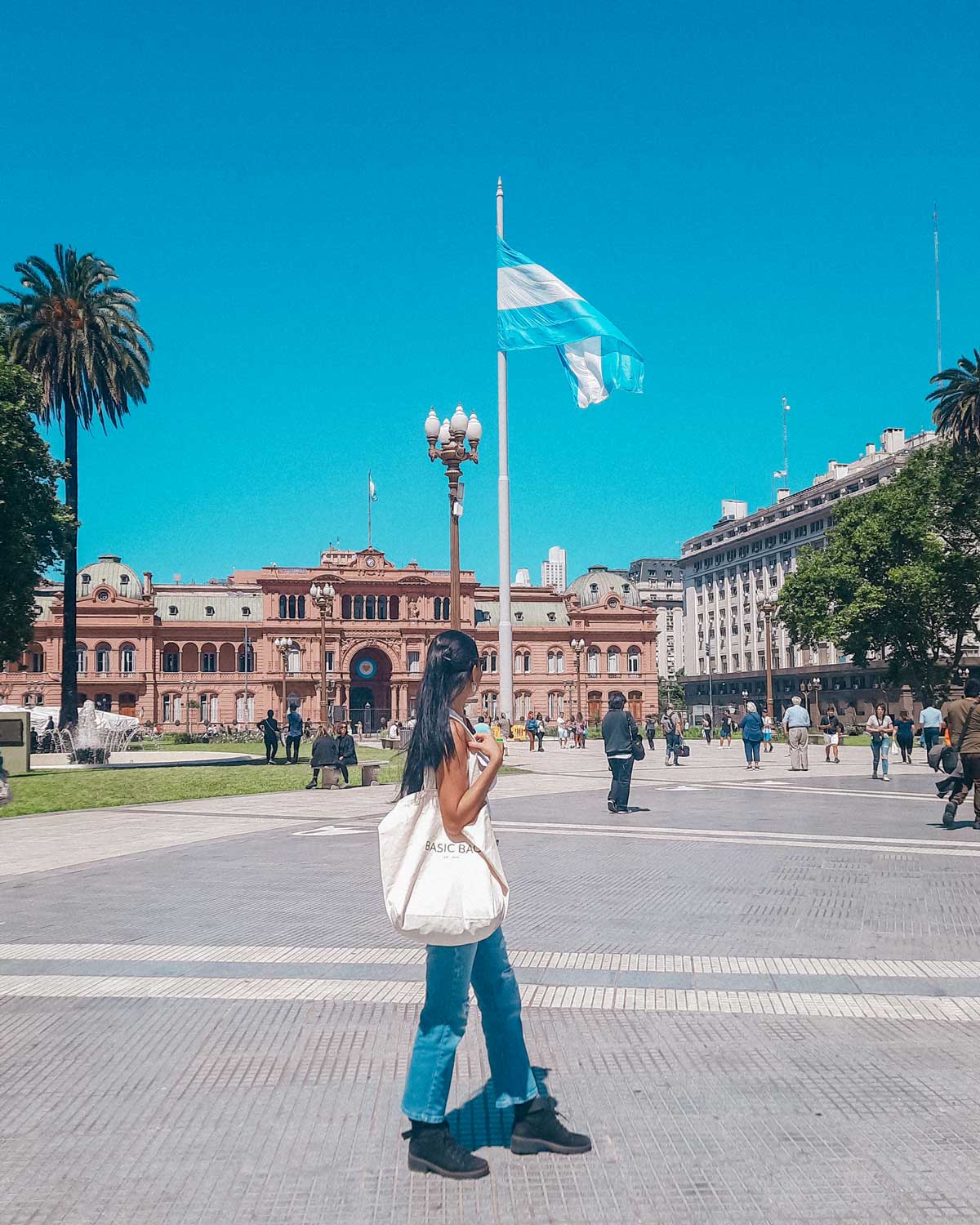 This image shows a woman walking in front of the Casa Rosada, the presidential palace of Argentina, located in Plaza de Mayo, Buenos Aires. The iconic pink building is visible in the background, with a large Argentine flag waving in the middle of the plaza. The woman, holding a bag labeled "BASIC BAG," walks across the plaza, surrounded by other pedestrians and palm trees under a bright, clear sky.