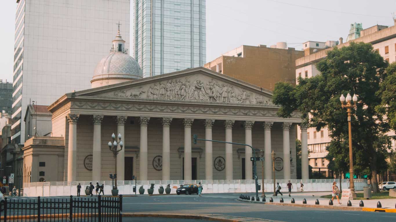 This image shows the Buenos Aires Metropolitan Cathedral, featuring its classical design with a grand neoclassical facade, complete with large columns and detailed bas-relief sculptures along the pediment. The dome of the cathedral rises behind the front structure, while modern buildings surround it, creating a juxtaposition of historical and contemporary architecture. A few pedestrians can be seen walking near the cathedral, contributing to the urban setting of this significant religious and historical site in the city.