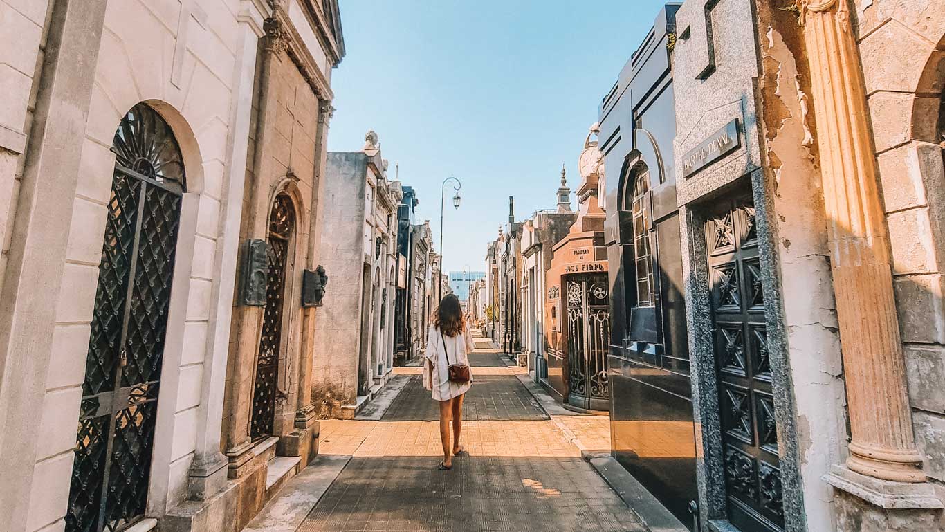 This image shows a woman walking through the narrow, ornate pathways of the Recoleta Cemetery in Buenos Aires, Argentina. The tombs on either side feature intricate designs, ranging from classic to modern, with various carvings and ironwork. The peaceful, solemn atmosphere of the cemetery is captured, as the woman, dressed in casual clothes, quietly explores this famous and historical resting place, known for its impressive mausoleums and architectural beauty.