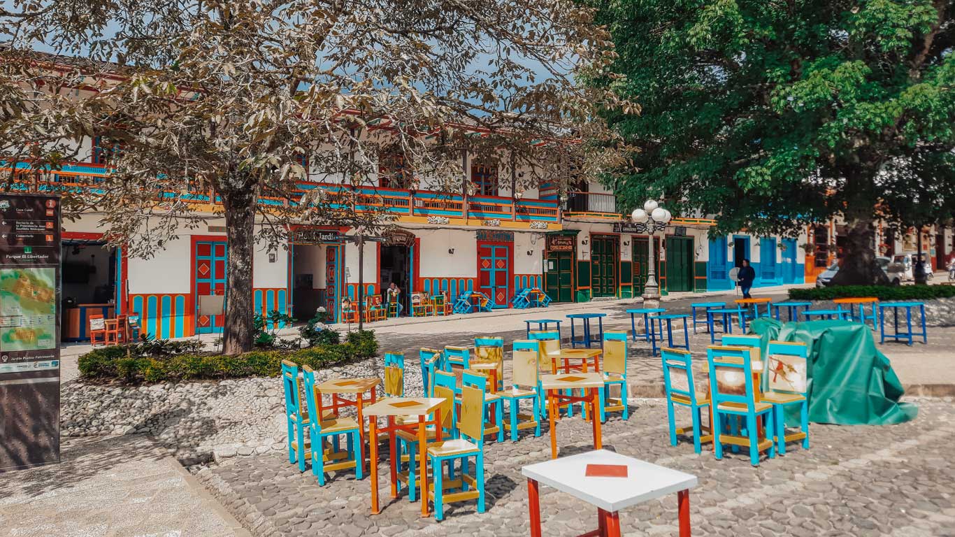 This image captures a colorful town square with bright blue, yellow, and red chairs and tables arranged on a cobblestone area, shaded by a large tree. In the background, a traditional building with vibrant accents and a sign that reads "Hotel Jardín" adds charm to the scene. The building features a second-story balcony with flowers, and the square appears peaceful and inviting, with a few people scattered about. The atmosphere reflects the quaint and lively nature of this picturesque Colombian town.