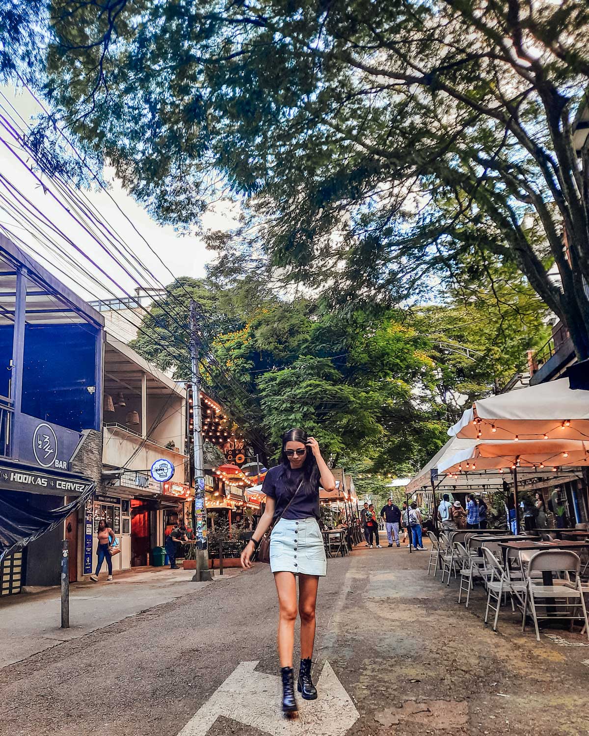 A woman in a black top and light blue denim skirt walks confidently down a street lined with outdoor cafes and shops. The scene is set in a lively urban area with greenery, string lights, and a relaxed atmosphere, likely capturing a moment in a trendy neighborhood. The arrow on the road beneath her points in her direction, adding a sense of motion to the image.