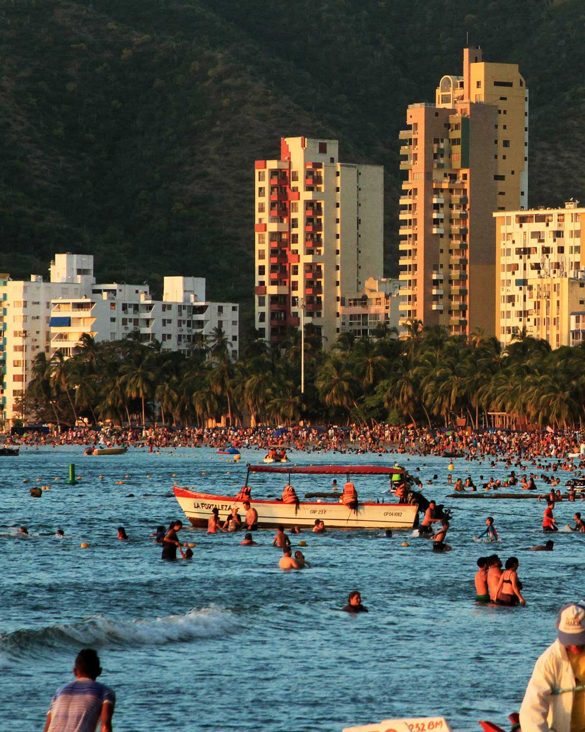 El Rodadero Beach, packed with people enjoying the sea, is one of the best places to stay in Santa Marta. In the background, there are tall buildings with lush green mountains behind them, creating a contrast with the palm trees along the coast.