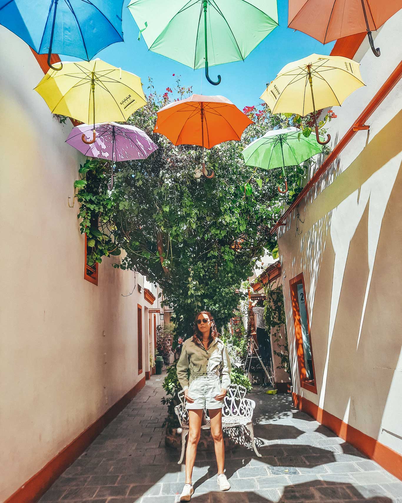 This image shows a woman standing in a colorful alley at Galería Salar French in Buenos Aires, Argentina. Above her, a series of vibrant umbrellas in shades of blue, yellow, green, and orange hang from strings, creating a playful and artistic canopy. The narrow alley is lined with white walls accented with red trim, and greenery adds to the lively atmosphere. The woman, dressed in light-colored clothing and sunglasses, stands in the shade, enjoying the unique and whimsical setting. This spot offers a fun photo opportunity for visitors during their 3 days in Buenos Aires.