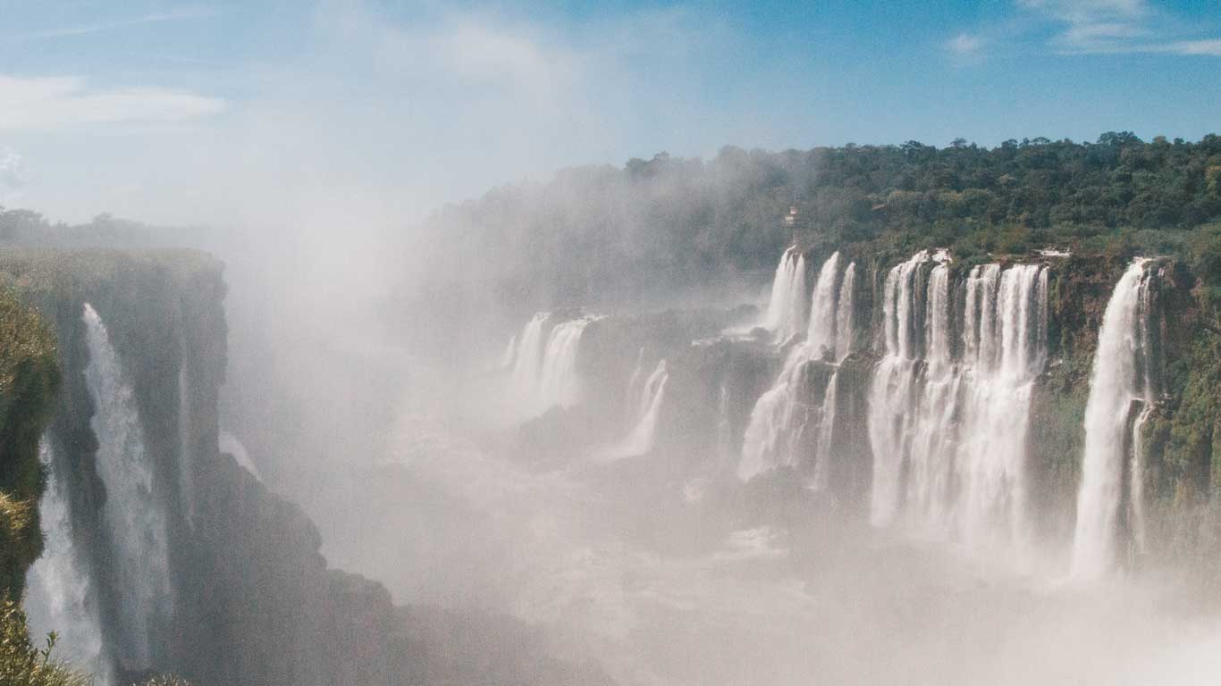 This image captures a misty view of Iguazu Falls, showcasing the impressive series of waterfalls cascading over lush cliffs. The dense spray from the powerful water flow partially obscures the scene, creating a dramatic and ethereal atmosphere against a backdrop of forested landscape and blue sky.