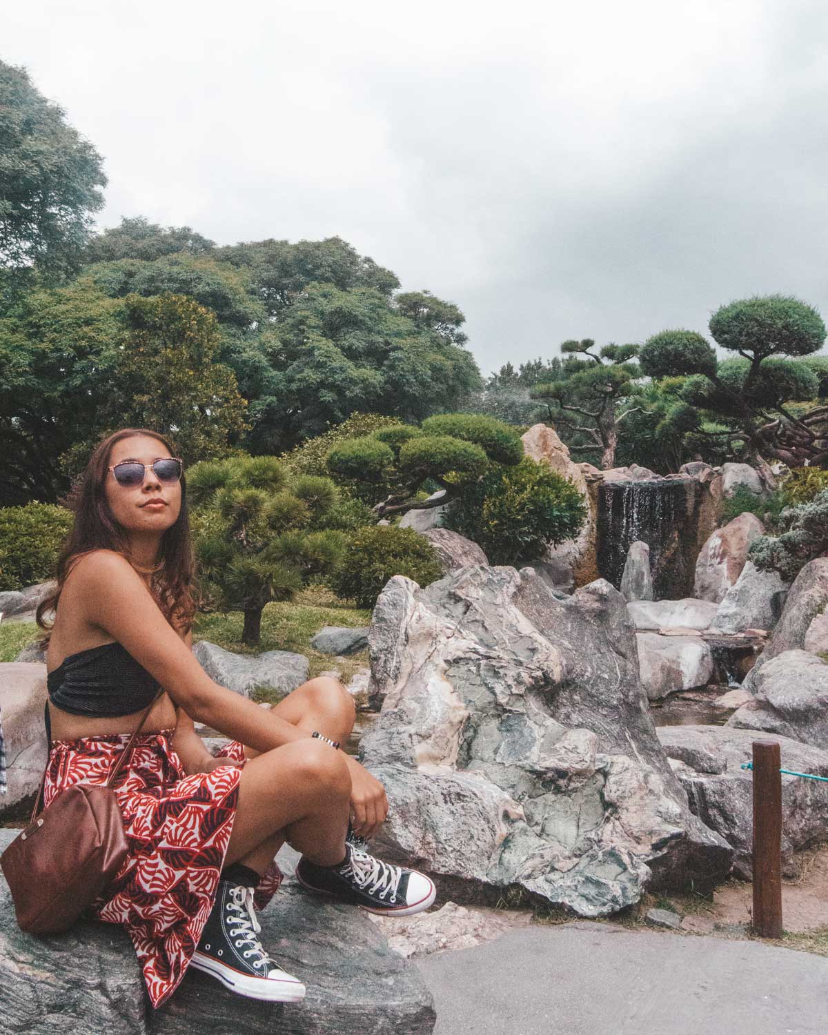 This image shows a young woman sitting on a large rock in the Jardín Japonés, a Japanese garden in Buenos Aires, Argentina. She is wearing sunglasses, a patterned red skirt, and black Converse sneakers, posing in a relaxed manner. In the background, there is a peaceful waterfall surrounded by meticulously pruned trees and large boulders, adding to the tranquil ambiance of the garden. The sky is overcast, further enhancing the calm and serene setting.