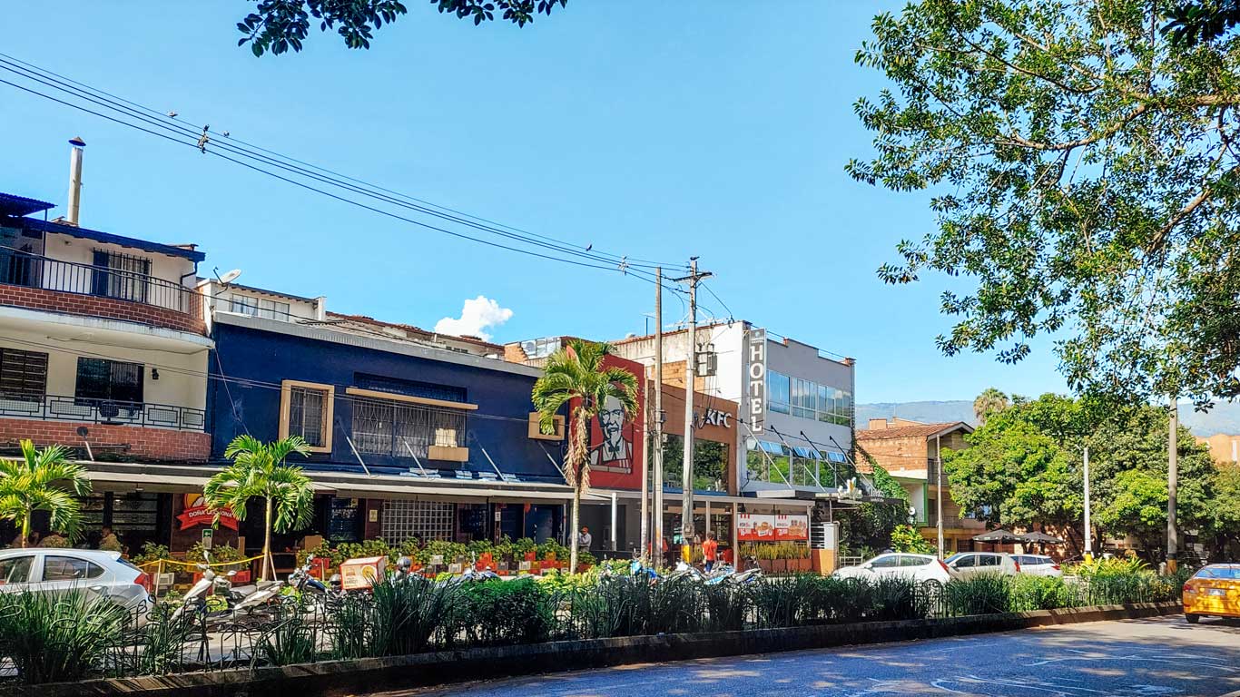 The image shows a street scene in a neighborhood with a mix of small businesses and greenery. A KFC restaurant is prominently visible, with a hotel sign next to it, and other buildings with various architectural styles. The sky is clear and blue, adding to the calm atmosphere of the area.