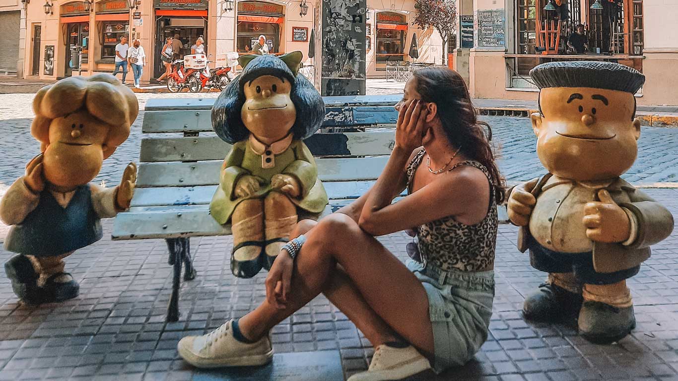 This image shows a woman sitting beside statues of the beloved comic strip character Mafalda and her friends, Manolito and Susanita, at the famous Mafalda bench in San Telmo, Buenos Aires. The woman, dressed casually, is playfully covering her face while seated near the iconic figures on the bench. The background includes a charming cobblestone street with local shops and pedestrians, capturing the lively and nostalgic atmosphere of this popular tourist spot, often visited during a Buenos Aires 3-day itinerary.