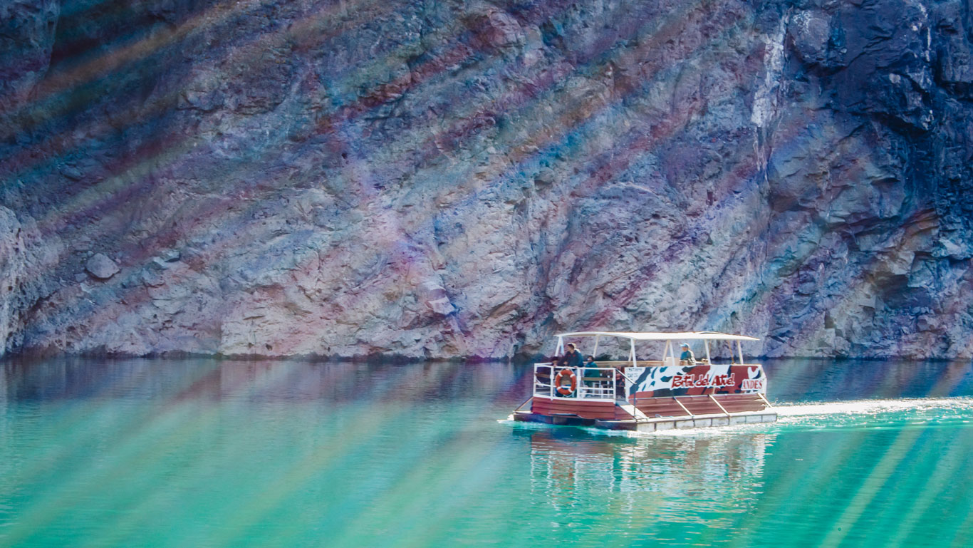 The image shows a tour boat called 'Rafa del Atuel Andes' sailing through turquoise waters on a lake surrounded by rocky walls. Sunlight creates a diagonal ray effect over the scene. The surroundings are mountainous, and the boat carries a group of people enjoying the natural landscape, typical of the San Rafael region in Mendoza.