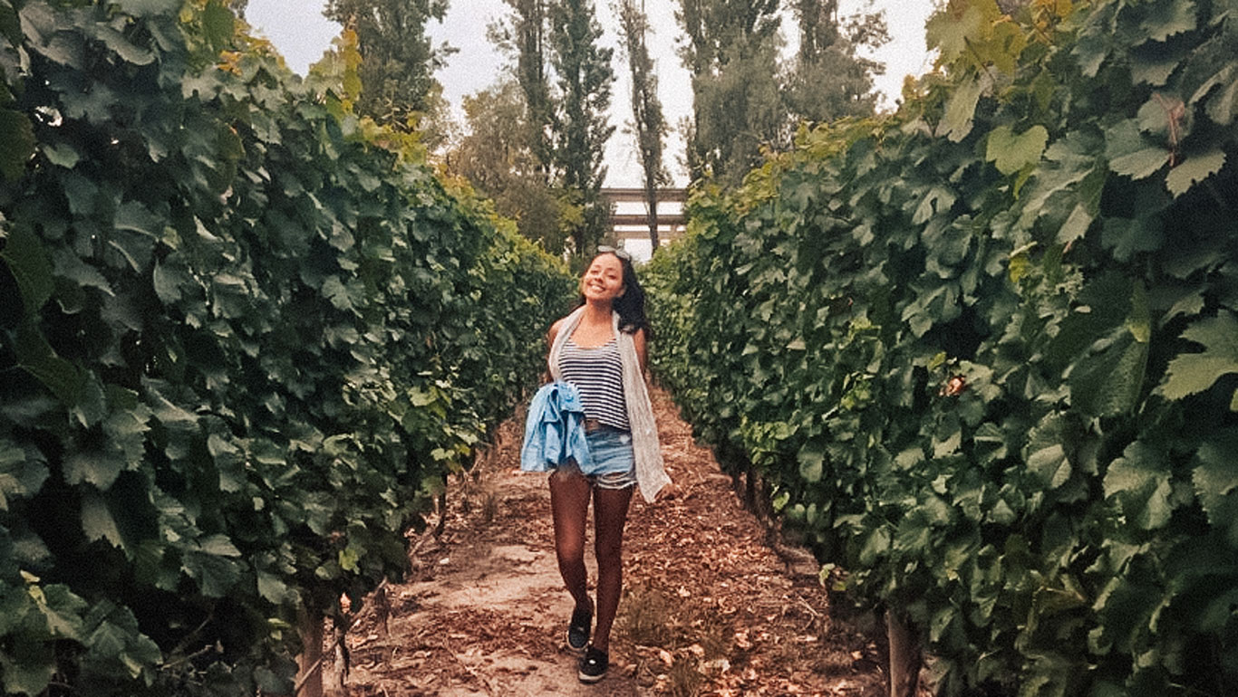 The image shows a woman happily walking through vineyards, surrounded by tall rows of green vines. She is wearing a casual summer outfit with a striped shirt and shorts, while holding a light jacket in her hand. The landscape reflects a natural and serene setting, typical of the wine regions of Mendoza.