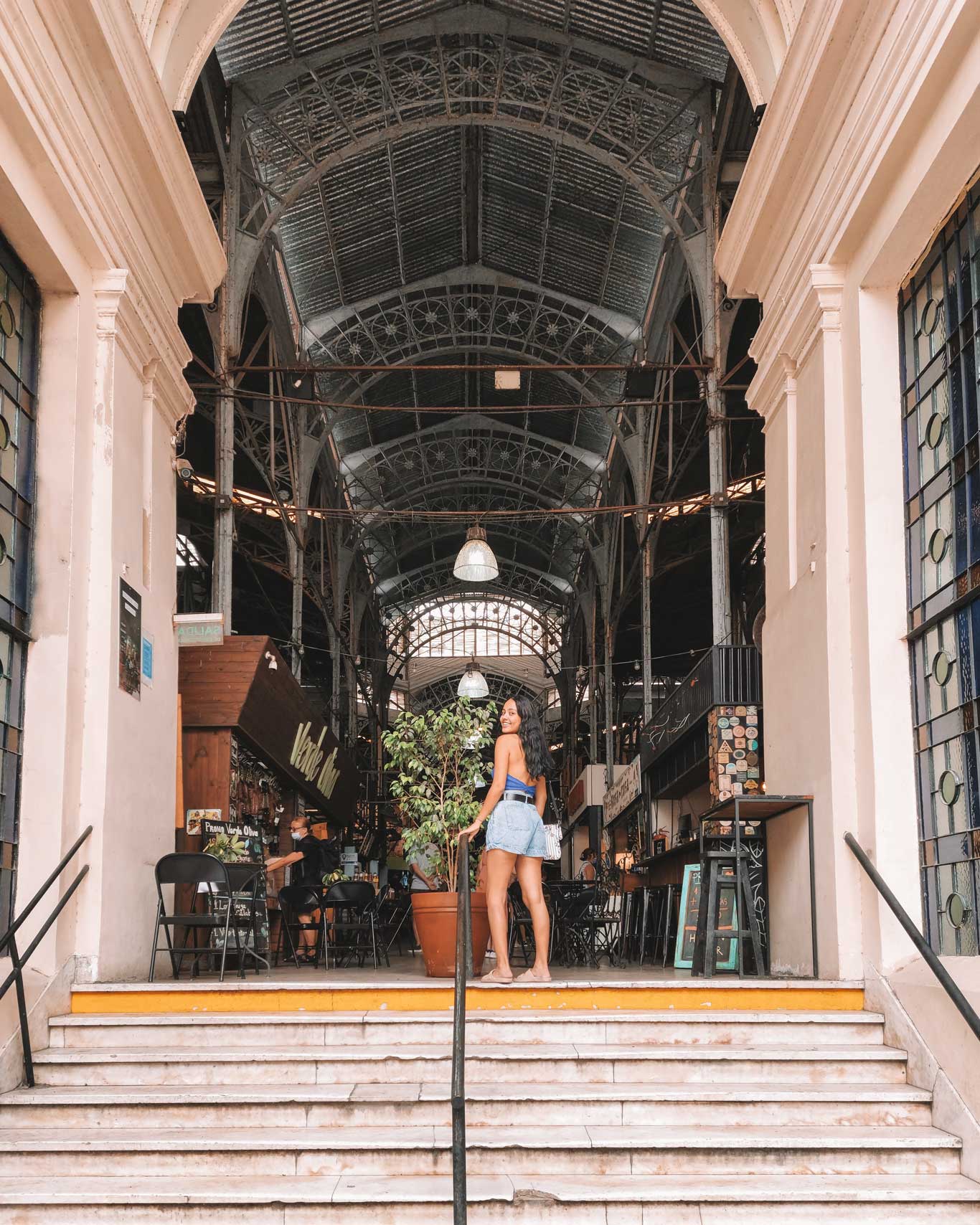 This image shows a woman standing at the entrance of the Mercado de San Telmo, a historic market in Buenos Aires, Argentina. She is posing on a set of stairs, smiling and looking back at the camera. The market's interior features high, ornate metal ceilings and a variety of shops and eateries visible inside. The architectural details and lively atmosphere make this market a popular destination for anyone exploring San Telmo during their 3 days in Buenos Aires.