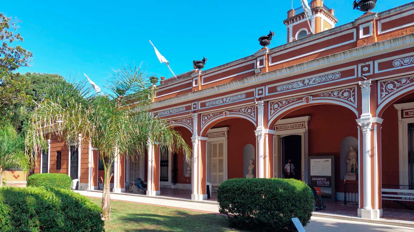 This image shows the exterior of the Museo Histórico de Buenos Aires, featuring its classic colonial architecture with ornate white columns and archways against a terracotta-colored facade. Palm trees and neatly trimmed bushes frame the entrance, adding a tropical touch to the scene. The bright blue sky overhead enhances the vibrant, sunny day. This museum is a popular stop for visitors exploring cultural landmarks during their 3 days in Buenos Aires.