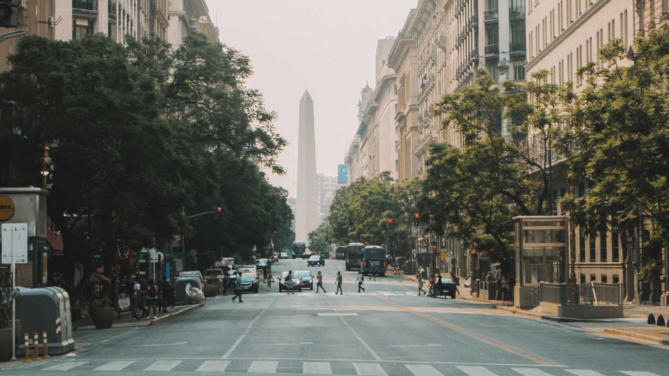 This image captures a view down a wide avenue in Buenos Aires, Argentina, with the iconic Obelisk (Obelisco de Buenos Aires) visible in the distance. The street is lined with trees and grand buildings, and a few pedestrians are crossing at a crosswalk. Vehicles are scattered along the road, contributing to the bustling urban atmosphere. The Obelisk, a well-known city landmark, stands tall in the center, framed by the surrounding architecture and greenery.