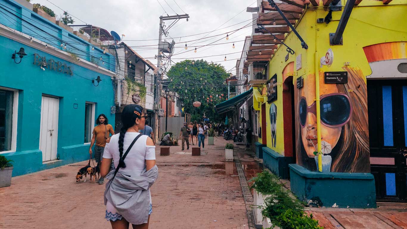 A pedestrian street in the historic downtown of Santa Marta, with colorful buildings on one side blue and on the other yellow with vibrant murals. People walk along the street under a festoon of hanging lights, and there is a relaxed, artistic feel to the place.