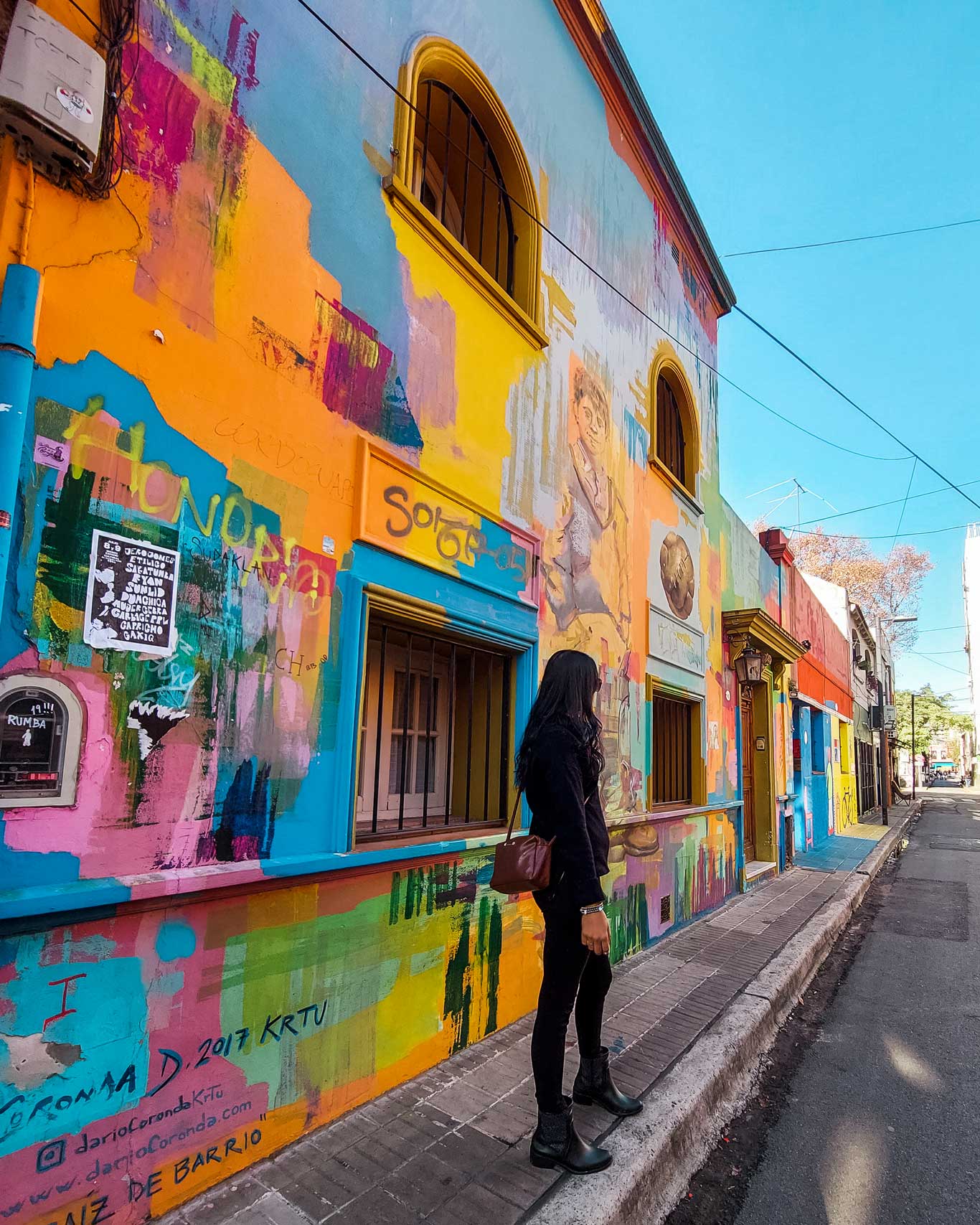 This image shows a colorful street scene in the Palermo neighborhood of Buenos Aires, Argentina. A woman dressed in black stands on the sidewalk, admiring a vibrant building covered in bold, abstract street art. The mural features splashes of orange, yellow, blue, and pink, with various artistic elements and graffiti, including portraits and text. The bright hues of the artwork contrast against the clear blue sky, adding to the energetic and creative vibe of the street.