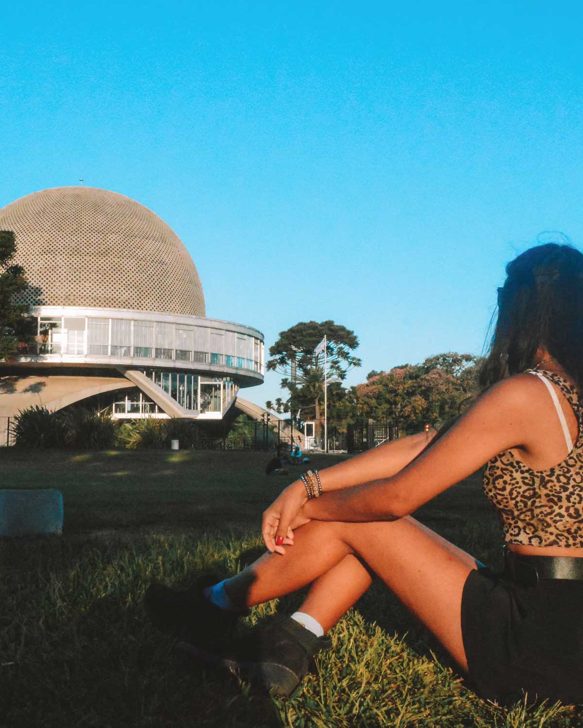 This image shows a woman sitting on the grass in front of the Planetario Galileo Galilei, a famous planetarium in Buenos Aires, Argentina. The woman is wearing a leopard-print top and black shorts, enjoying the sunny day while gazing toward the planetarium's distinct dome-shaped structure. The clear blue sky and green lawn create a peaceful, outdoor setting, with the planetarium's modern architecture standing out prominently in the background.