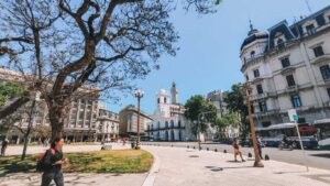 This image shows Plaza de Mayo in Buenos Aires, Argentina, featuring historical buildings and a bright, sunny atmosphere. In the center is the Cabildo building with its iconic white facade and tower. A tree in the foreground stretches its branches, while pedestrians walk along the sidewalk. Surrounding buildings exhibit classic European-style architecture.