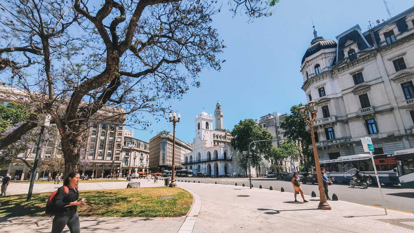 This image shows Plaza de Mayo in Buenos Aires, Argentina, featuring historical buildings and a bright, sunny atmosphere. In the center is the Cabildo building with its iconic white facade and tower. A tree in the foreground stretches its branches, while pedestrians walk along the sidewalk. Surrounding buildings exhibit classic European-style architecture.