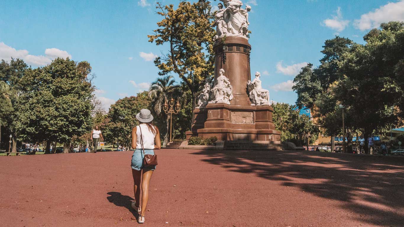 This image shows a woman walking towards a large monument in Plaza Francia, located in the Recoleta neighborhood of Buenos Aires, Argentina. The statue is surrounded by lush green trees, and the bright day highlights the park's vibrant and peaceful atmosphere. The woman, dressed casually in a hat and shorts, strolls through the park, emphasizing the leisurely and open space of this popular outdoor area. The monument itself features detailed sculptures, adding to the historic and artistic charm of the plaza.