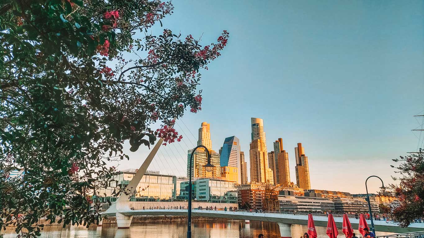 This image features the Puente de la Mujer (Woman's Bridge) in the Puerto Madero district of Buenos Aires, Argentina. The bridge's sleek, modern design spans the water, with a line of people crossing it. In the background, the city skyline is bathed in warm sunset light, highlighting the tall glass and steel skyscrapers. The foreground includes blossoming trees and red umbrellas, creating a picturesque and lively scene of this contemporary urban area.