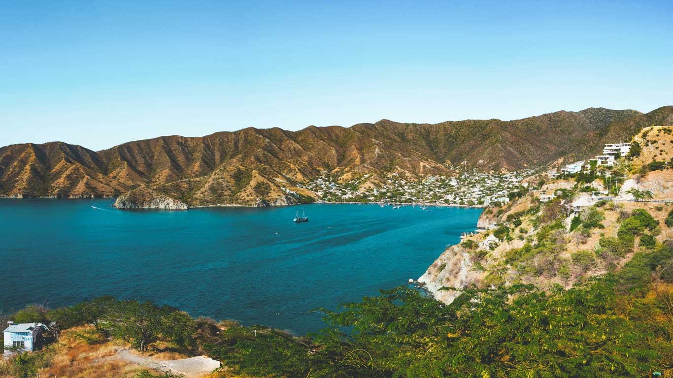 A tranquil Taganga bay with blue waters surrounded by green mountains. Small boats are seen in the distance, and the coast is dotted with houses and sparse vegetation.