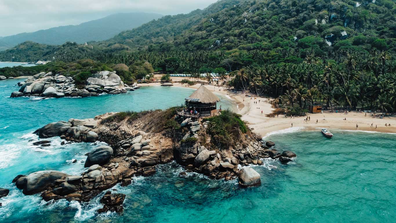 An aerial view of a small rocky peninsula in Tayrona Park, with a cabin on top surrounded by crystal-clear water. In the background, a white sand beach is surrounded by dense vegetation and green mountains. The park is also an excellent choice for where to stay in Santa Marta.