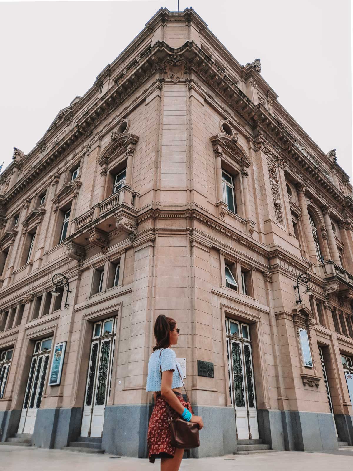 This image captures a woman standing in front of the Teatro Colón in Buenos Aires, Argentina, admiring its grand, neoclassical architecture. The theater's ornate stone facade is adorned with intricate details and large windows, with corner balconies and decorative elements adding to its stately appearance. The woman, dressed casually with a crossbody bag, stands in contrast to the historic and imposing building.