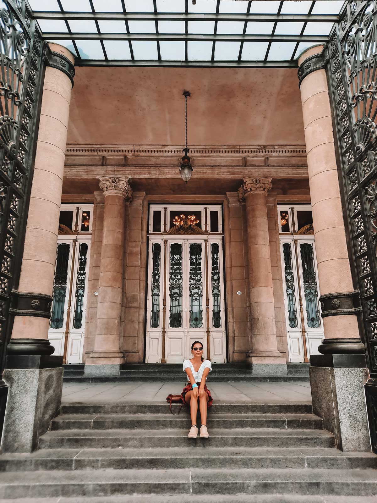 This image shows the entrance of the Teatro Colón in Buenos Aires, Argentina, featuring grand stone pillars and ornate, wrought-iron doors. A woman is seated on the steps in front of the entrance, wearing sunglasses and casual attire. The intricate architectural details of the theater’s facade, along with the glass roof above, emphasize its historic and elegant design.