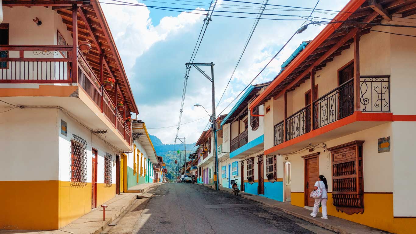 This image shows a quiet street in Jardín lined with colorful buildings with vibrant orange, blue, yellow and white facades. The architecture includes wooden balconies and decorative railings, adding charm to the traditional style. A woman in white is walking along the sidewalk, carrying a bag, while mountains are visible in the background under a blue sky with scattered clouds. The scene reflects a peaceful and picturesque atmosphere in the center of the village, a great place where to stay in Jardín.