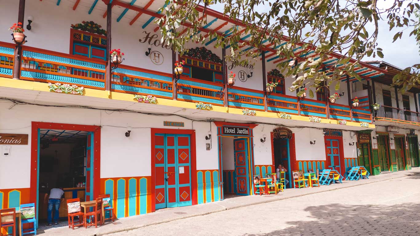 This image shows a brightly colored hotel facade with vibrant red, blue, yellow, and green accents. The building has a traditional colonial-style design with a balcony adorned with flower pots and wooden decorative elements. The sign on the building reads "Hotel Jardín," indicating it as a hotel in the area. Several small tables and chairs are set up outside, adding to the inviting and charming atmosphere of this picturesque town street.