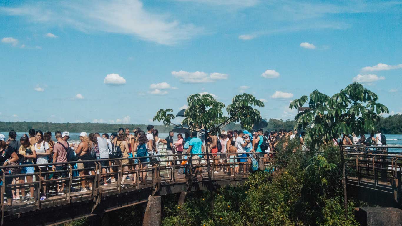 The image shows a crowded walkway near Iguazu Falls, with numerous tourists gathered to enjoy the view. The wooden platform, surrounded by greenery, provides a scenic lookout over the falls under a bright, partly cloudy sky, highlighting the popularity of this natural attraction.