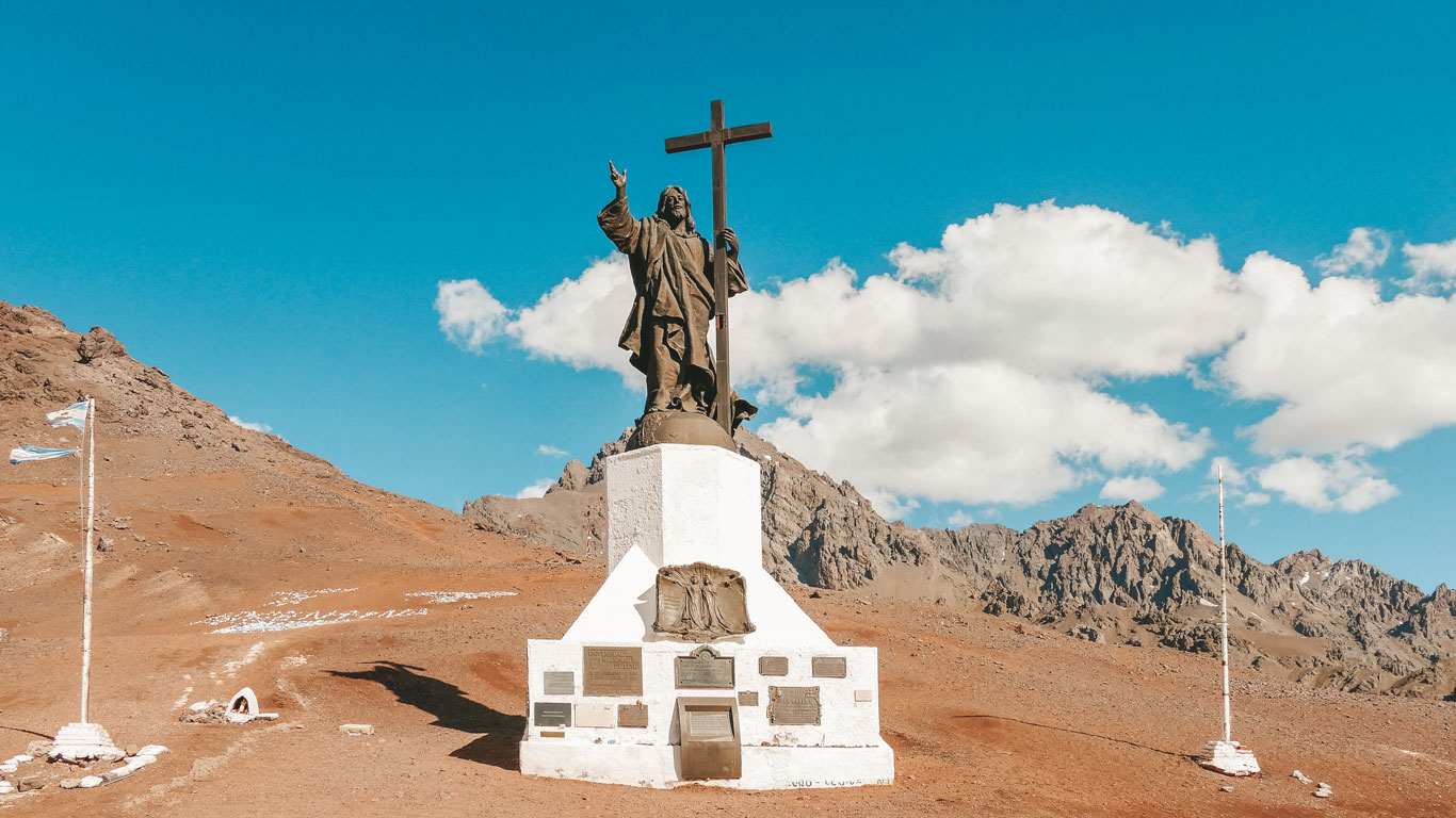 An imposing statue of Christ holding a cross, set on a white pedestal with commemorative plaques, surrounded by an arid mountain landscape in the Andes. In the background, rocky peaks and a clear blue sky with a few clouds add depth to the scene. Two Argentine flags wave on either side, emphasizing the historical and spiritual significance of the location.