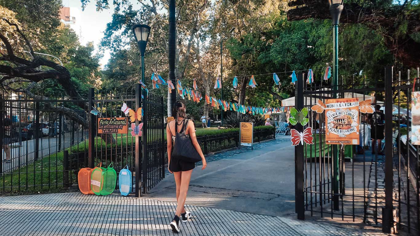 This image captures a vibrant entrance to Plaza Belgrano in Buenos Aires, decorated with colorful flags and butterfly motifs for an artisanal fair. A woman walks through the gates, emphasizing the welcoming and community-focused atmosphere of the space. 