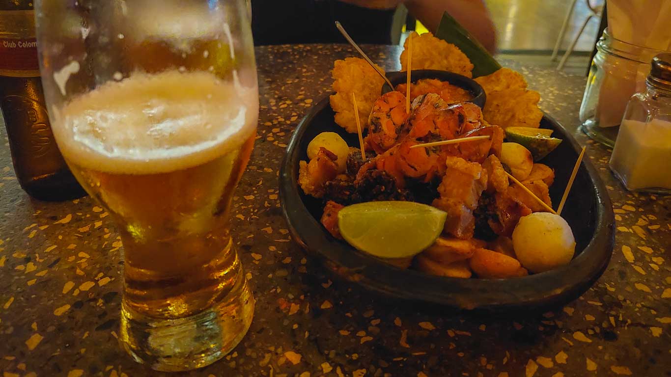 This image shows a close-up of a healthy meal accompanied by a glass of beer at Mamasina, one of the best restaurants in Medellín. The dish features a variety of grilled meats and fried sides, including skewered pieces, lemon wedges, and possibly plantain or cassava chips. The golden beer and the rustic, dimly lit atmosphere add to the inviting dining experience.