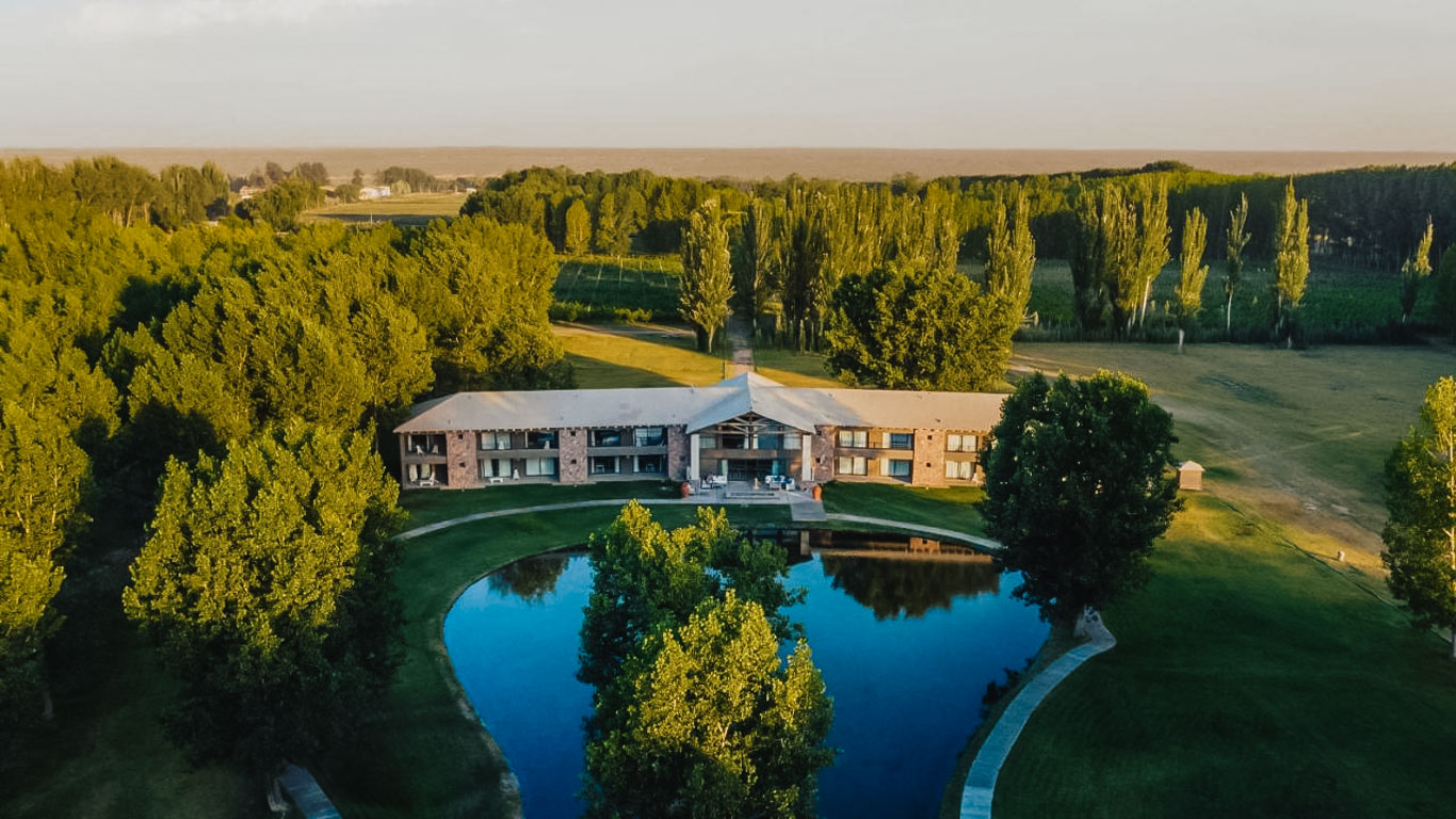 This image features a serene aerial view of a countryside property surrounded by lush green trees and open fields. At the center, a two-story brick building with large windows and a modern design overlooks a reflective pond bordered by manicured lawns. The expansive, natural setting conveys tranquility and rural charm.