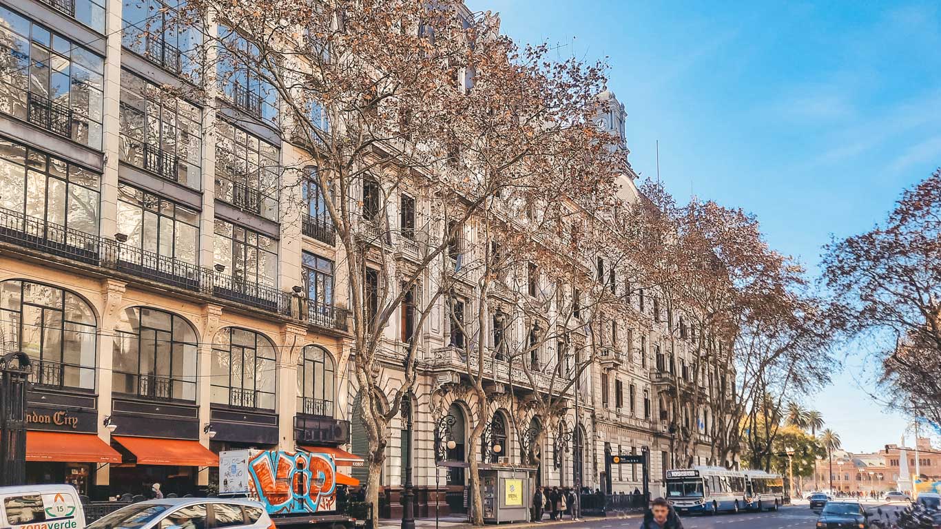 This image captures the vibrant downtown area of Buenos Aires, showcasing elegant European-style architecture with large windows and intricate details. Leafless trees line the street, adding character to the urban setting, while bustling cafes and shops highlight the lively atmosphere.