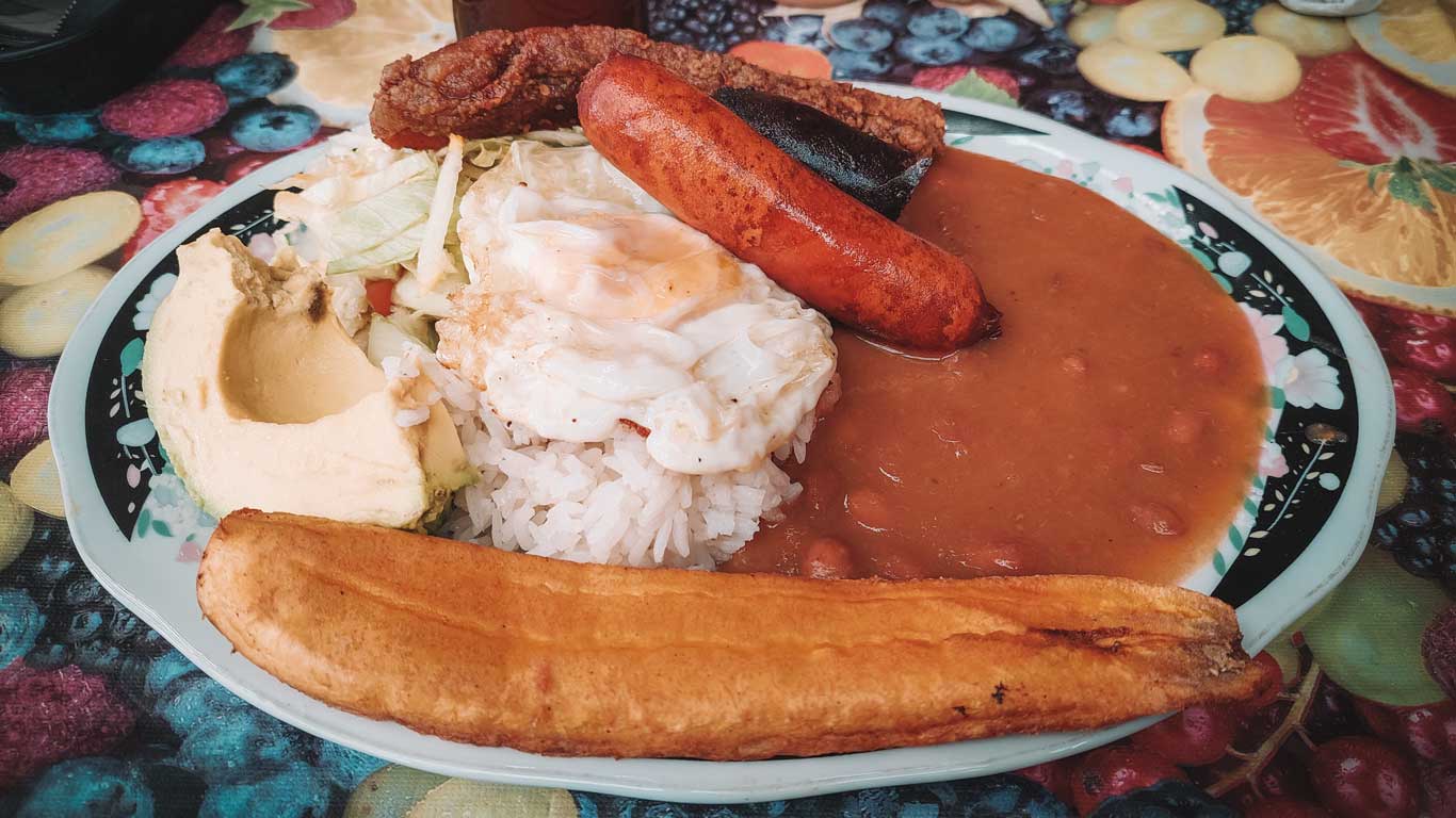 This image showcases a traditional Colombian meal, likely a bandeja paisa, featuring white rice topped with a fried egg, red beans in a rich sauce, chorizo sausage, crispy pork belly (chicharrón), fried plantain, and fresh avocado. The vibrant plate is set against a colorful, fruit-patterned tablecloth, emphasizing the hearty and flavorful nature of the dish.